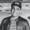Black-and-white portrait of a smiling team member from Digital Marketing Toolkit wearing a baseball cap, striped shirt, and dark leather jacket, standing outdoors against a softly blurred natural background and facing the camera with a relaxed, professional expression.
